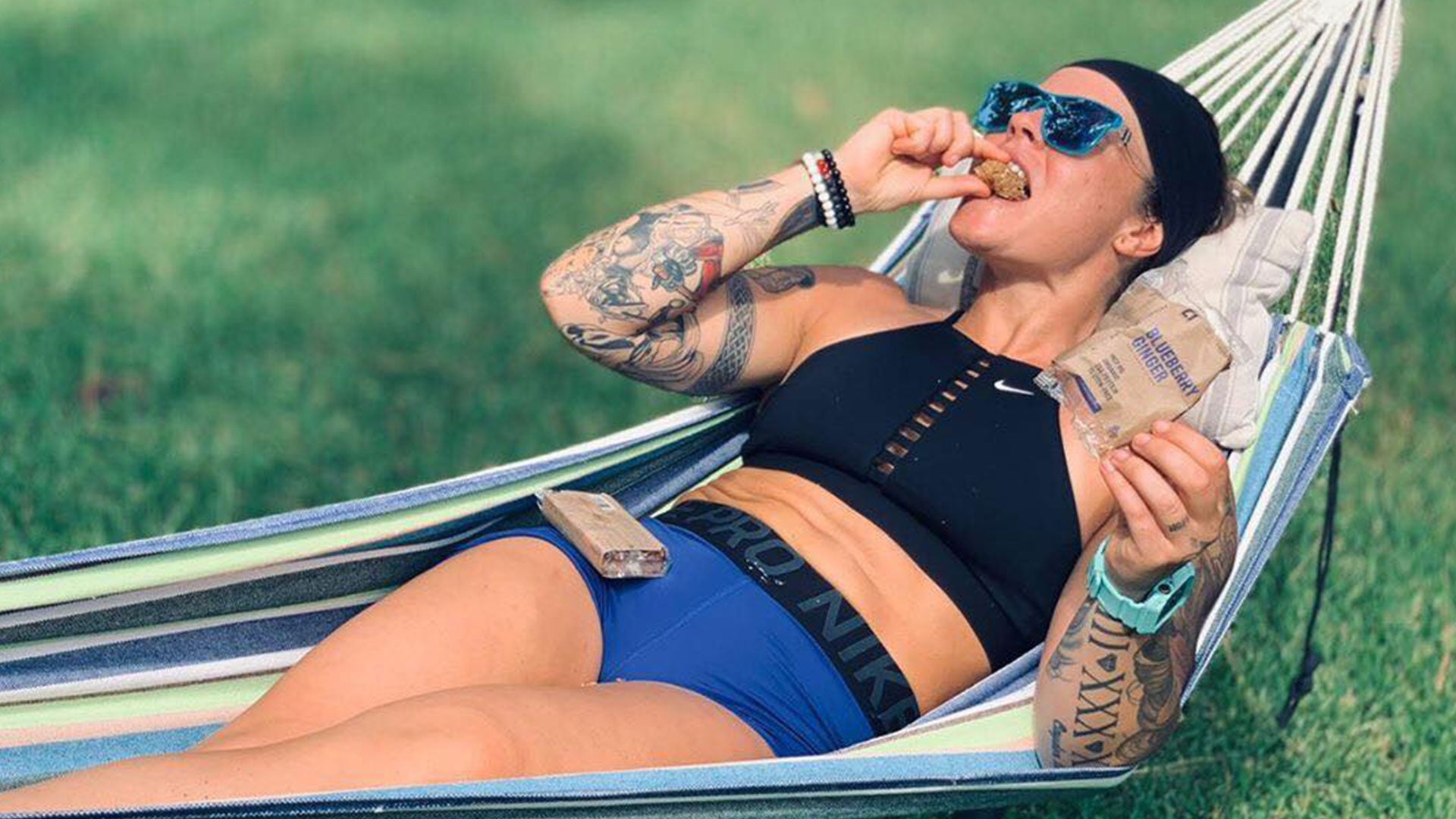 Woman lounging in a hammock wearing a black sports bra and blue shorts, eating a C1 bar.