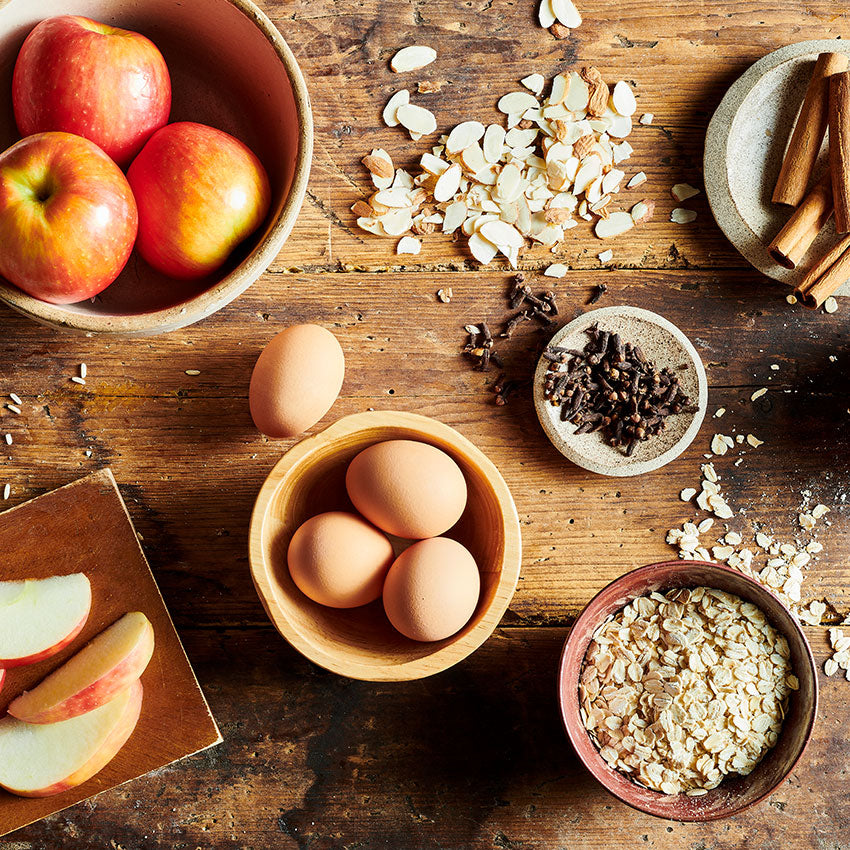 Wooden table with bowls of apples, eggs, oats, and other ingredients.